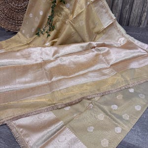 May include: A shimmering, golden-toned fabric with a delicate floral pattern and a fringed border. The fabric is draped, showcasing its texture and sheen. The overall aesthetic suggests a luxurious textile, possibly for apparel or home decor.