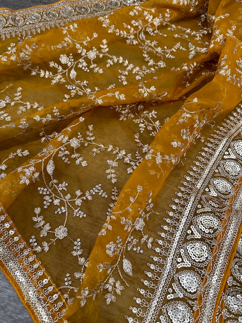 May include: A mustard yellow sheer fabric with a silver floral embroidery pattern. The fabric has a silver sequined border.