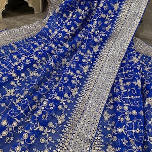 May include: Royal blue fabric with intricate floral embroidery and silver sequins. The fabric is draped, showcasing the detailed design and texture. The edges are lined with a border of silver sequins.