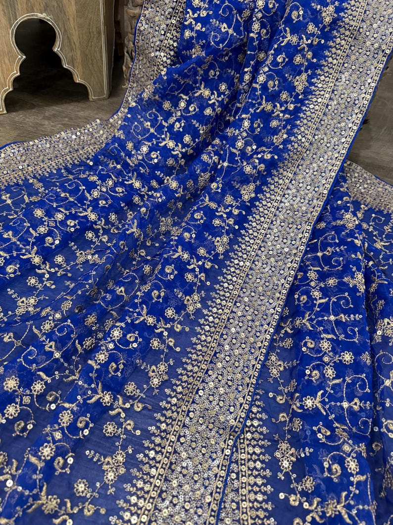 May include: Royal blue fabric with intricate floral embroidery and silver sequins. The fabric is draped, showcasing the detailed design and texture. The edges are lined with a border of silver sequins.