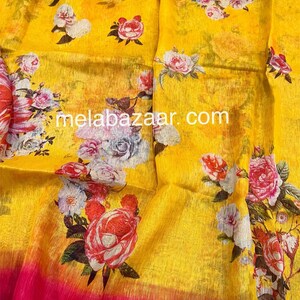 May include: Yellow linen saree with a pink border and floral print. The print features roses, daisies, and other flowers in shades of pink, white, and red. The saree has a subtle ombre effect, with the yellow fading to a lighter shade at the bottom.