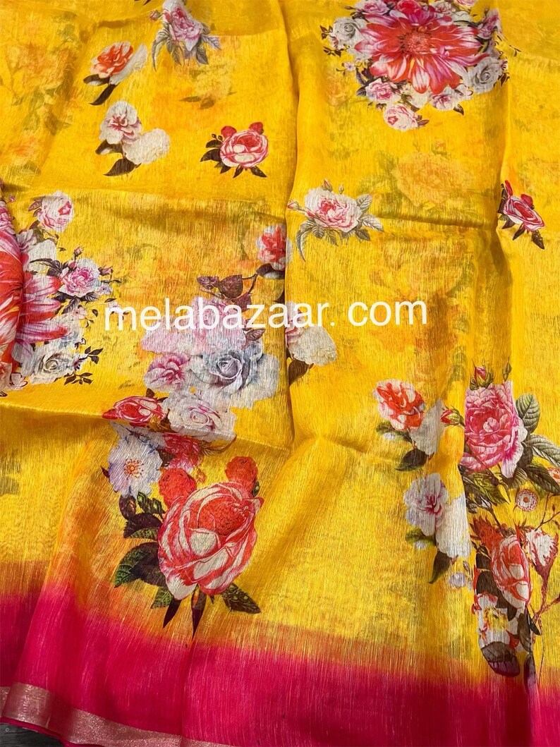 May include: Yellow linen saree with a pink border and floral print. The print features roses, daisies, and other flowers in shades of pink, white, and red. The saree has a subtle ombre effect, with the yellow fading to a lighter shade at the bottom.