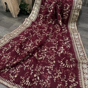 Könnte beinhalten: Ein weinrotes Textil mit aufwendiger goldener Blumenstickerei. Der Stoff hat einen detaillierten Rand mit goldenen Verzierungen. Das Textil ist über einem Weidenkorb drapiert, wodurch seine Textur und sein Design hervorgehoben werden. Die Gesamtästhetik ist elegant und aufwendig.