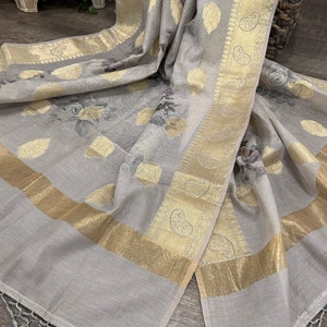 May include: A gray and gold floral patterned saree with a gold border and tassels. The saree is made of a soft, lightweight fabric and has a delicate floral design.