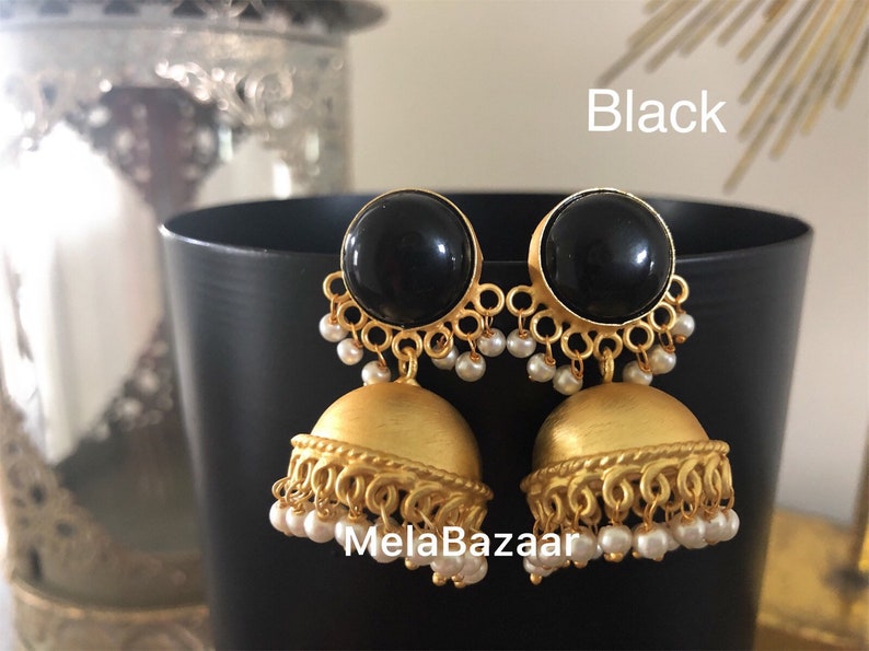 May include: A pair of black and gold-toned earrings. The earrings feature a large, round black stone at the top, with a gold-colored filigree design and small white beads. The bottom portion is a gold-colored bell shape. The word "Black" is visible.