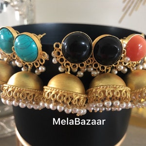 May include: A collection of gold-toned earrings with various gemstone tops, including turquoise, black, and coral. The earrings feature a bell-shaped design with intricate detailing and small pearl accents. The brand name "MelaBazaar" is visible.