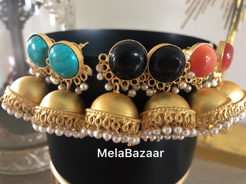May include: A collection of gold-toned earrings with various gemstone tops, including turquoise, black, and coral. The earrings feature a bell-shaped design with intricate detailing and small pearl accents. The brand name "MelaBazaar" is visible.