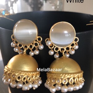 May include: A pair of gold-colored earrings with a white, round top and a bell-shaped bottom. The earrings have a pearl-like bead design and the text "MelaBazaar" and "White".