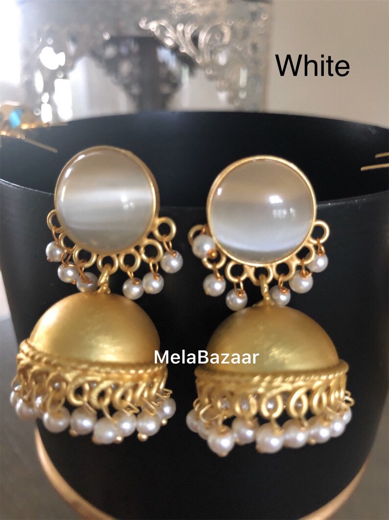 May include: A pair of gold-colored earrings with a white, round top and a bell-shaped bottom. The earrings have a pearl-like bead design and the text "MelaBazaar" and "White".