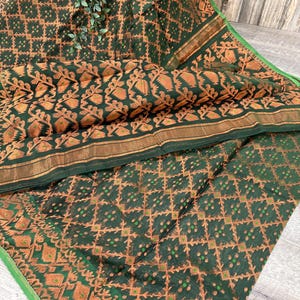 May include: A dark green textile with an intricate pattern in shades of orange and gold. The fabric features a repeating geometric design with floral accents. A thin gold border and a green edge frame the textile, which is draped on a light-colored surface.
