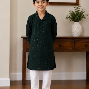May include: A dark green long-sleeved tunic with a collar and a buttoned placket, featuring a subtle shimmering texture throughout the fabric. The tunic is paired with plain white trousers and simple black slip-on shoes.