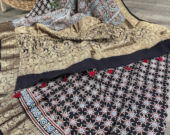 May include: A draped textile featuring intricate patterns in black, gold, and various colors. The fabric displays floral and geometric designs, with a gold border and red tassel accents. The textile appears to be a saree or similar garment.