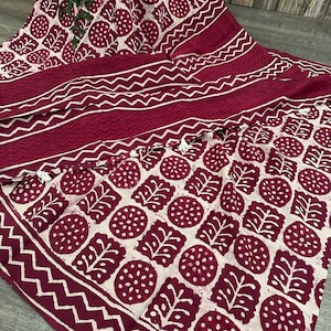 K&ouml;nnte beinhalten: Bordeaux- und cremefarbener Stoff mit einem sich wiederholenden Muster aus Bl&auml;ttern und Kreisen. Der Stoff hat einen Rand mit Zickzack-Design und wei&szlig;en Quasten. Der Stoff ist drapiert und zeigt sein Design.