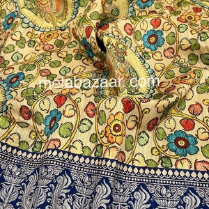 May include: A cream-colored fabric with a floral pattern in shades of blue, green, yellow, and red. The fabric has a blue border with a silver-colored design of peacocks and floral motifs. The text "melabazar.com" is visible in the center of the fabric.