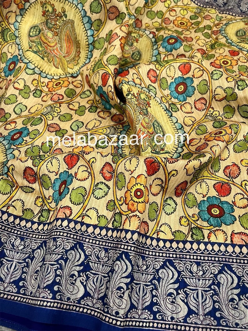 May include: A cream-colored fabric with a floral pattern in shades of blue, green, yellow, and red. The fabric has a blue border with a silver-colored design of peacocks and floral motifs. The text "melabazar.com" is visible in the center of the fabric.