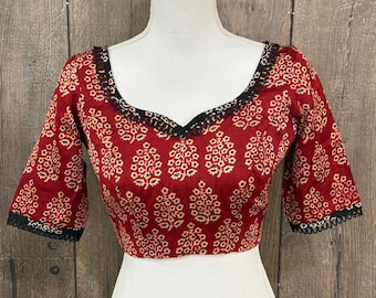 May include: A red blouse with a floral pattern and black trim. The blouse has a V-neckline and three-quarter sleeves. The floral pattern is white and beige. The trim is a black, scalloped design. The blouse is displayed on a white mannequin.