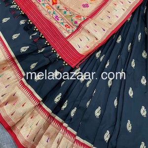May include: A black silk saree with a gold and red border. The saree has a paisley pattern and is decorated with gold tassels. The saree is folded in half and the border is visible. The saree is a traditional Indian garment.