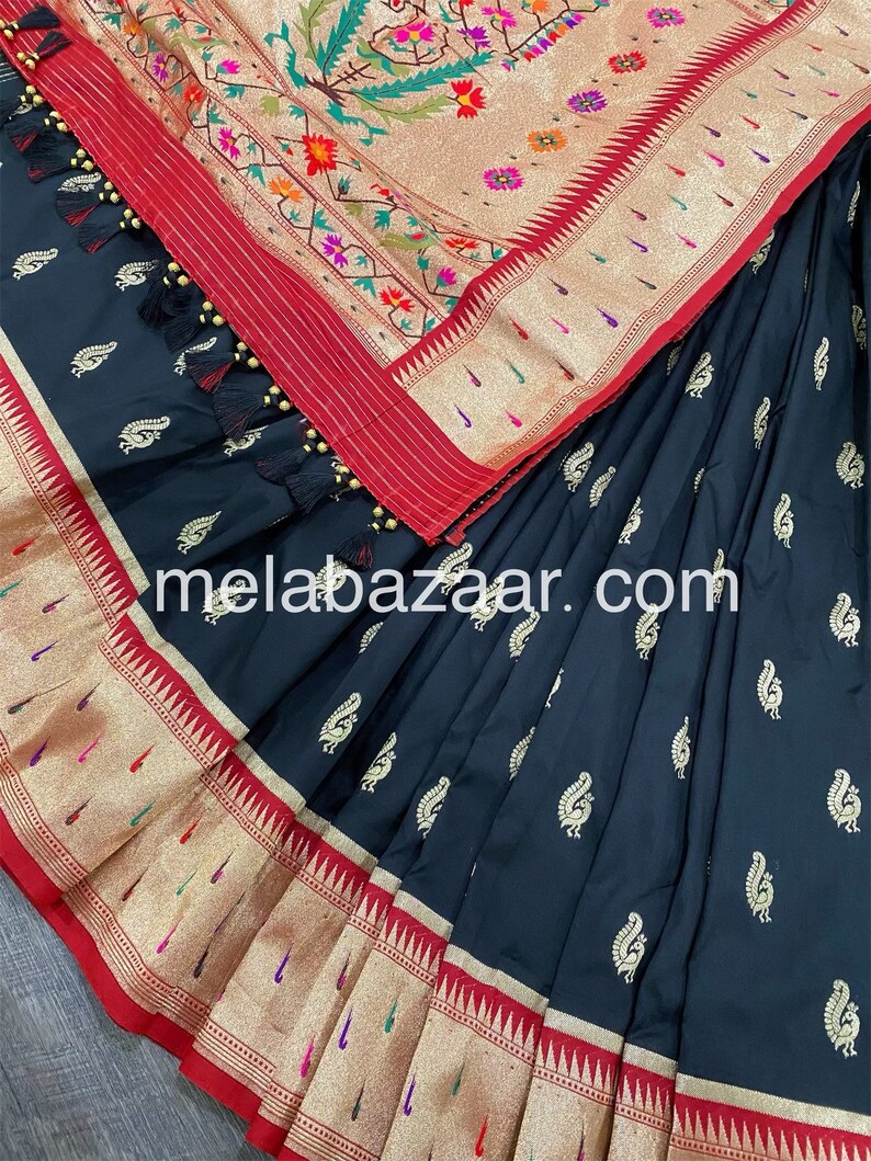 May include: A black silk saree with a gold and red border. The saree has a paisley pattern and is decorated with gold tassels. The saree is folded in half and the border is visible. The saree is a traditional Indian garment.