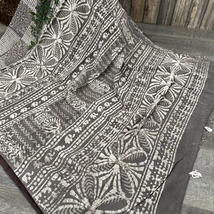 Puede incluir: Una tela de algod&oacute;n gris con un patr&oacute;n batik blanco de flores y dise&ntilde;os geom&eacute;tricos. La tela tiene un borde con flecos.