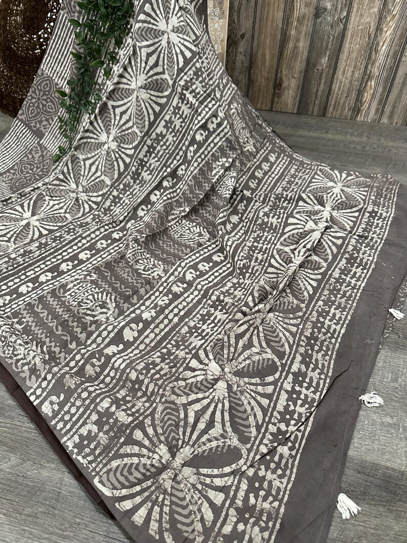 Puede incluir: Una tela de algod&oacute;n gris con un patr&oacute;n batik blanco de flores y dise&ntilde;os geom&eacute;tricos. La tela tiene un borde con flecos.