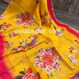 May include: Yellow linen saree with a floral print in shades of pink, white, and green. The saree has a pink border and tassels. The text "melabazar.com" is printed on the saree.