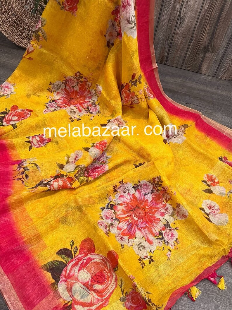 May include: Yellow linen saree with a floral print in shades of pink, white, and green. The saree has a pink border and tassels. The text "melabazar.com" is printed on the saree.