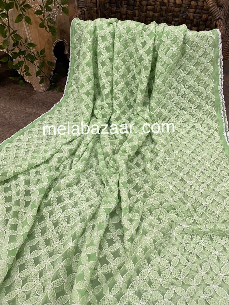 May include: A light green chiffon fabric with white embroidered floral designs. The fabric has a delicate, sheer texture and a subtle sheen.