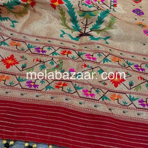 May include: A gold and red silk saree with a floral pattern and a black tassel border. The saree has a red border with gold stripes and a gold and red floral pattern. The saree is decorated with gold thread embroidery. The saree is a traditional Indian garment worn by women.
