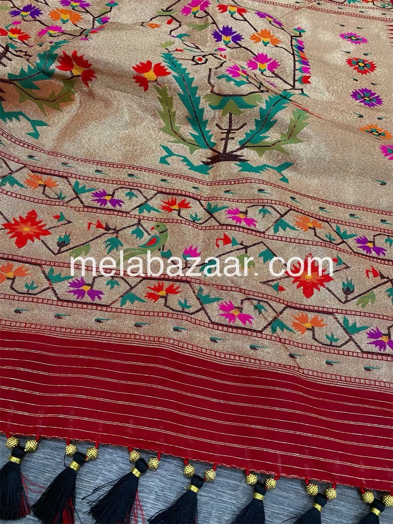May include: A gold and red silk saree with a floral pattern and a black tassel border. The saree has a red border with gold stripes and a gold and red floral pattern. The saree is decorated with gold thread embroidery. The saree is a traditional Indian garment worn by women.