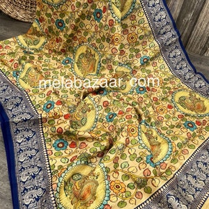 May include: A cream-colored saree with a blue border and a floral print featuring peacocks. The saree has a traditional Indian design and is made of a soft, lightweight fabric.