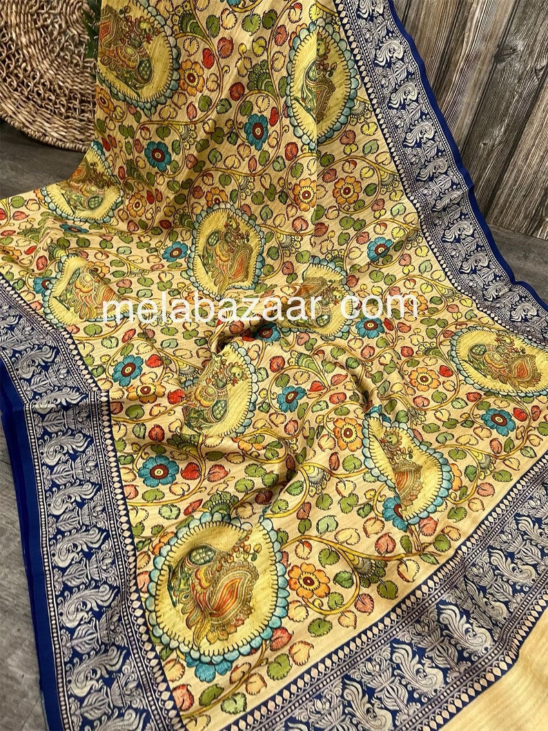 May include: A cream-colored saree with a blue border and a floral print featuring peacocks. The saree has a traditional Indian design and is made of a soft, lightweight fabric.