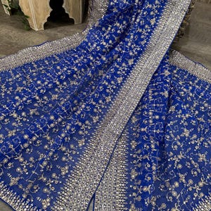 May include: A royal blue textile with intricate silver embroidery and sequin detailing. The fabric features a floral pattern and a wide border of sequins. The textile is draped over a woven basket and wooden surface.