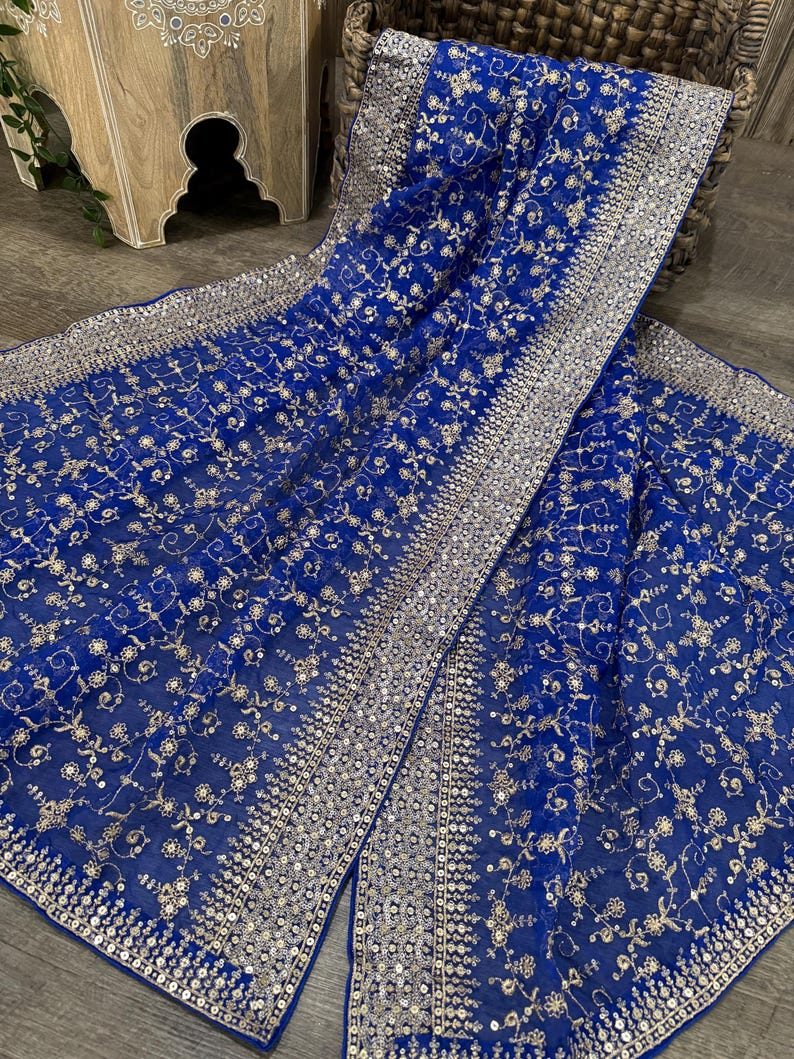 May include: A royal blue textile with intricate silver embroidery and sequin detailing. The fabric features a floral pattern and a wide border of sequins. The textile is draped over a woven basket and wooden surface.