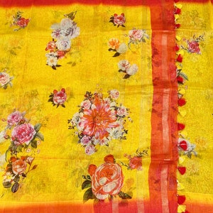 May include: Yellow linen saree with a floral print in shades of red, pink, and white. The saree has a red and gold border with tassels.