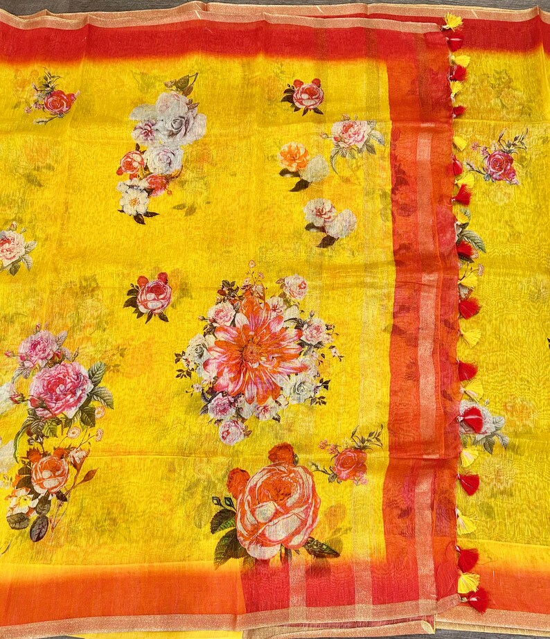 May include: Yellow linen saree with a floral print in shades of red, pink, and white. The saree has a red and gold border with tassels.