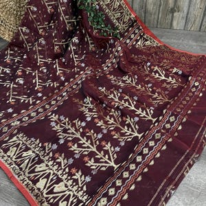 May include: A deep maroon saree featuring a repeating gold metallic tree motif across the fabric. The border displays intricate geometric patterns in gold, white, and orange threads, finished with a thin red trim along the outer edge.