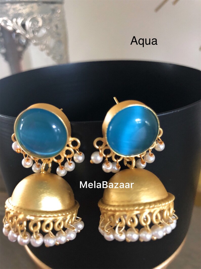 May include: Gold-toned earrings with a round, aqua-colored stone top and a bell-shaped bottom. The earrings are adorned with small, dangling white pearls. The word "Aqua" is visible in the image.