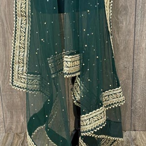 Puede incluir: Un dupatta verde oscuro transparente con bordados dorados y lentejuelas. El dupatta presenta intrincadas borduras doradas con un ribete de perlas. La tela está drapeada sobre un soporte negro, mostrando el diseño.