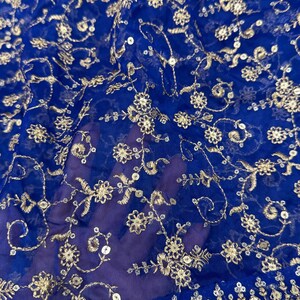 May include: Close-up of a sheer, royal blue fabric with intricate gold embroidery. The design features floral patterns and sequins, creating a delicate and ornate texture. The fabric is likely used for apparel or decorative purposes.