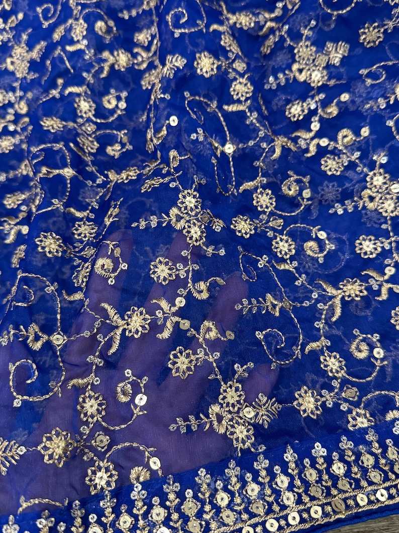 May include: Close-up of a sheer, royal blue fabric with intricate gold embroidery. The design features floral patterns and sequins, creating a delicate and ornate texture. The fabric is likely used for apparel or decorative purposes.