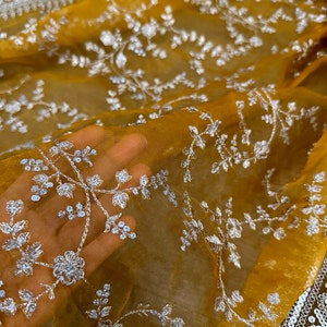 May include: A mustard yellow sheer fabric with white floral embroidery. The fabric is translucent and the embroidery is delicate.