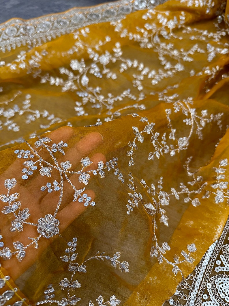 May include: A mustard yellow sheer fabric with white floral embroidery. The fabric is translucent and the embroidery is delicate.
