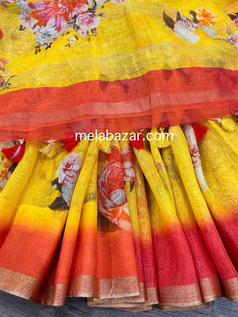May include: Yellow linen saree with a floral print and a red and orange ombre effect. The saree has a gold trim and tassels.