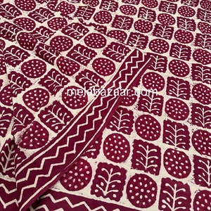 K&ouml;nnte beinhalten: Ein gefaltetes Textil mit einem sich wiederholenden Muster aus weinroten und wei&szlig;en botanischen Formen. Das Design umfasst Kreise, Quadrate und blattartige Formen. Ein Zickzack-Rand umrahmt den Rand. Die Website "melebazaar.com" ist sichtbar.