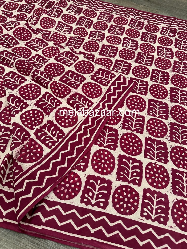 K&ouml;nnte beinhalten: Ein gefaltetes Textil mit einem sich wiederholenden Muster aus weinroten und wei&szlig;en botanischen Formen. Das Design umfasst Kreise, Quadrate und blattartige Formen. Ein Zickzack-Rand umrahmt den Rand. Die Website "melebazaar.com" ist sichtbar.