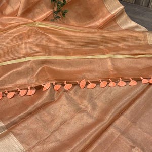 May include: Peach-colored sheer fabric with a subtle striped pattern and gold trim. The fabric is adorned with a row of peach-colored tassels along one edge. A sprig of green leaves adds a touch of natural contrast.