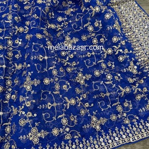 May include: Royal blue fabric with gold floral embroidery and sequin detailing. The fabric has a delicate, sheer quality, with the gold embroidery creating a rich, textured pattern. The border is embellished with sequins. The website address melabazaar.com is visible.