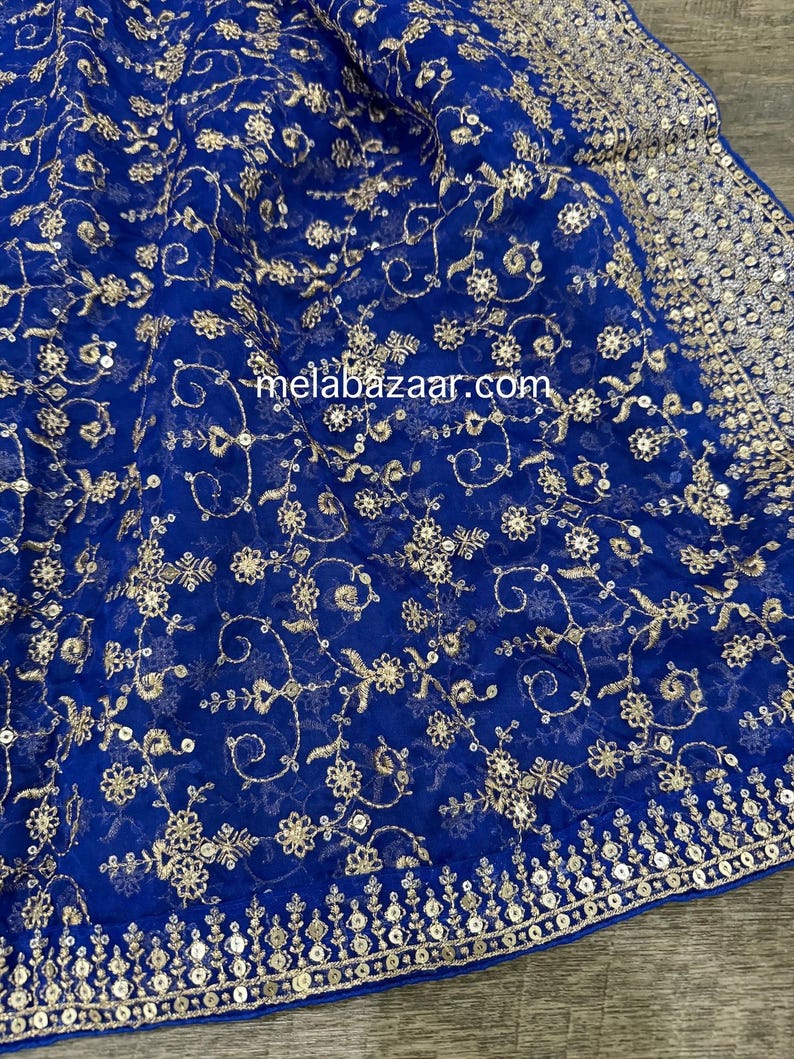 May include: Royal blue fabric with gold floral embroidery and sequin detailing. The fabric has a delicate, sheer quality, with the gold embroidery creating a rich, textured pattern. The border is embellished with sequins. The website address melabazaar.com is visible.
