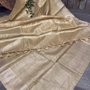 May include: A shimmering gold-toned saree featuring a translucent, lightweight fabric with a subtle metallic sheen. The garment includes horizontal cream-colored striped bands and a decorative border finished with small, matching gold-toned tassels.