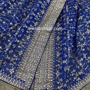 May include: Royal blue fabric with intricate gold floral embroidery and sequin detailing. The fabric is draped, showcasing the design and texture. The website melabazaar.com is visible on the fabric.